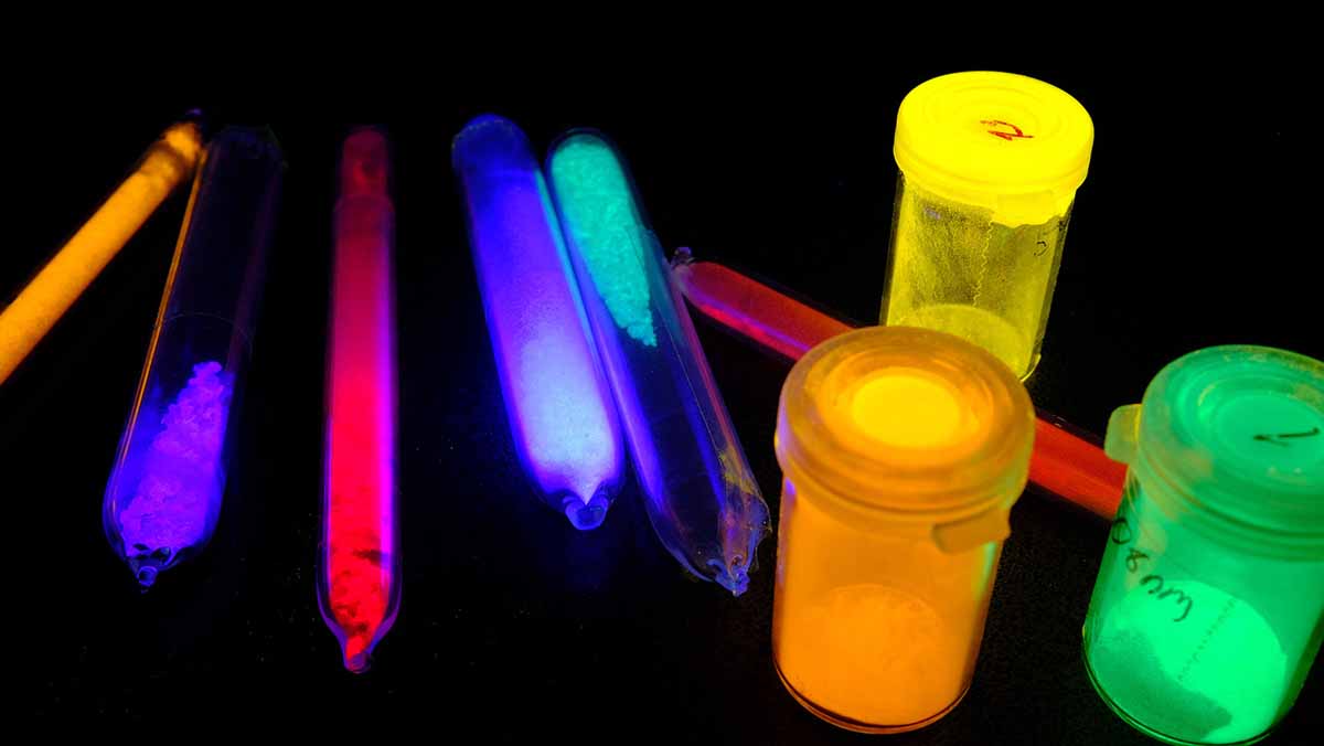 Probengläser gefüllt mit leuchtenden Teilchen in Neonfarben vor schwarzem Hintergrund