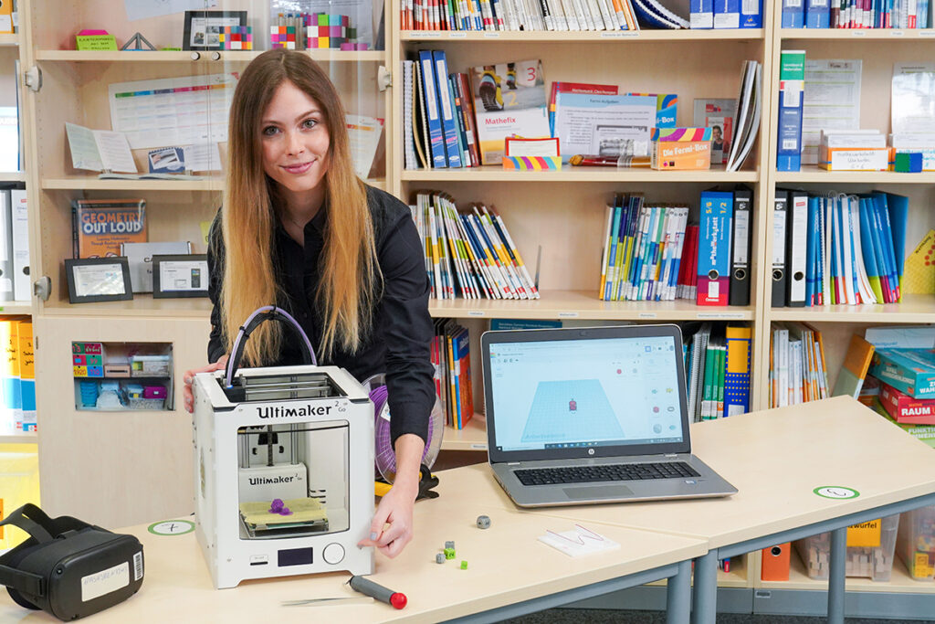 Frau steht hinter einem Tisch mit 3D-Drucker und Laptop