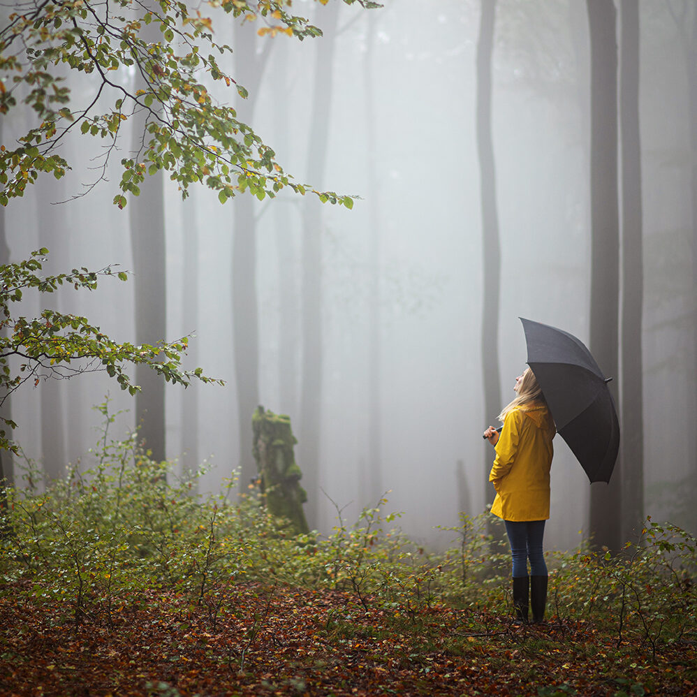 Eine Frau in gelbem Regenmantel steht in einem nebligen Wald