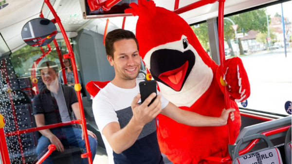 Mann mit Handy steht im Bus neben Vogel-Maskottchen