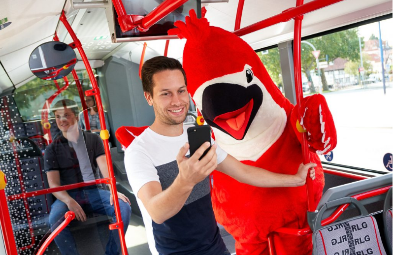 Mann mit Handy steht im Bus neben Vogel-Maskottchen