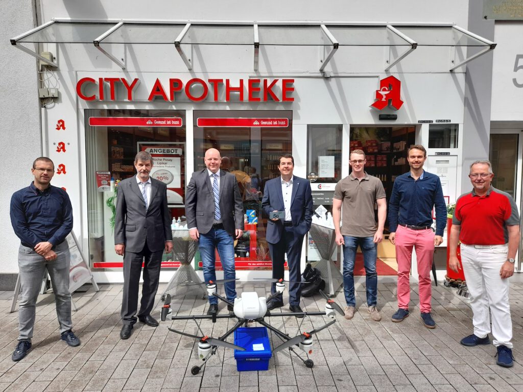 Sieben Personen stehen mit einer Paketdrohne vor einer Apotheke