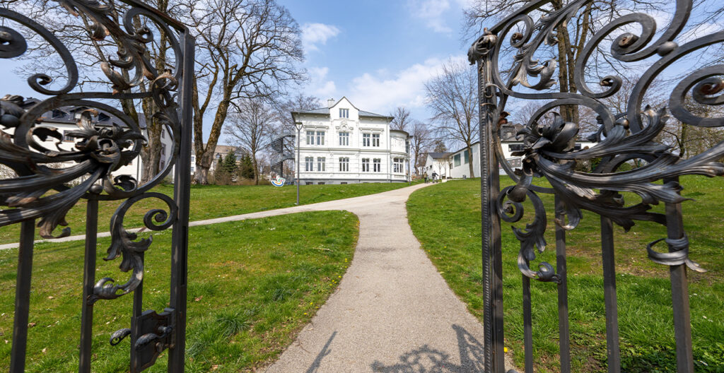 Blick durch ein geöffnetes eisernes Tor, das zu einem grünen Park mit einer Villa führt